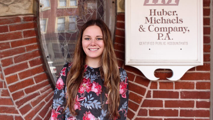 Young woman standing outside Huber Michaels & Company, P.A. accounting firm, smiling near brick wall.