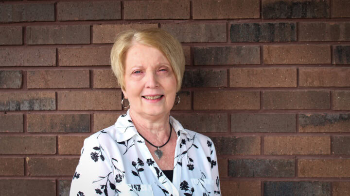 Elderly woman smiling outdoors against a brick wall, professional headshot for Huber Michaels & Company.