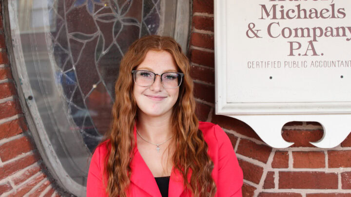 Young woman with red hair and glasses standing outside Huber Michaels & Company CPA firm in front of brick wall and business sign.